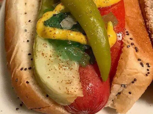 A close-up of a hot dog topped with mustard, pickles, and green peppers in a bun, sprinkled with spices.