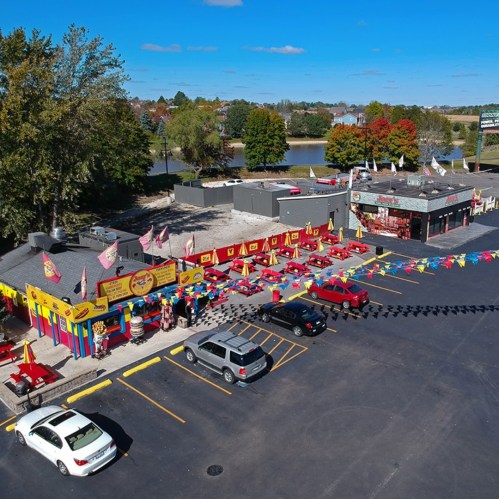Aerial view of a colorful restaurant with outdoor seating, surrounded by trees and a parking lot on a sunny day.