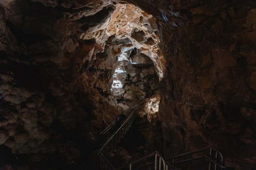 A dimly lit cave interior with rocky formations and a metal staircase leading deeper into the cavern.