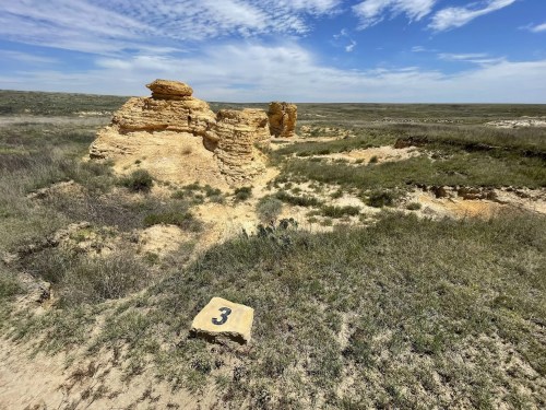 A rocky formation in a grassy landscape, with a numbered marker (3) in the foreground and a blue sky above.