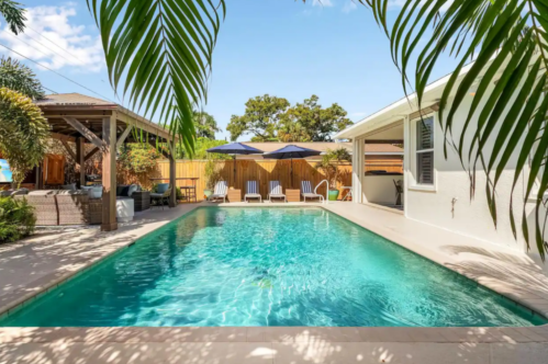 A serene backyard pool surrounded by lounge chairs and umbrellas, with tropical plants and a cozy seating area nearby.