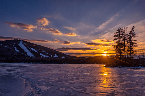 Sunset over a frozen lake with a mountain in the background and silhouetted trees, creating a serene winter landscape.