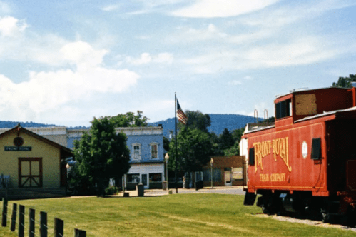A red caboose near a small town with a flag, surrounded by green grass and mountains in the background.