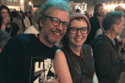 Two smiling people pose together at a lively event, surrounded by a crowd and holding drinks.