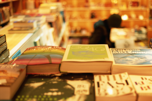 A stack of colorful books on a table, with a blurred figure browsing in the background of a cozy bookstore.
