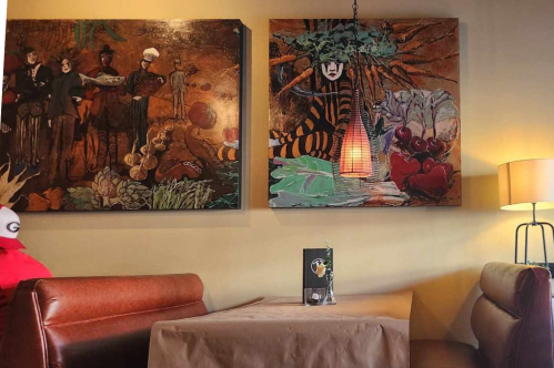 Cozy cafe interior featuring two colorful wall art pieces and a table set for dining. Warm lighting and seating visible.