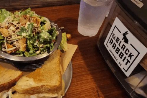 A bowl of salad next to a plate of grilled cheese sandwiches and a water glass, with a sign for The Black Rabbit.