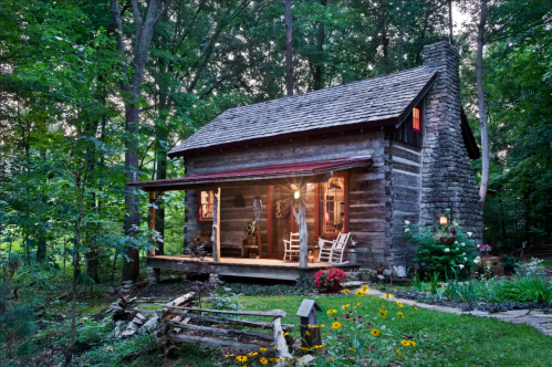 A rustic log cabin surrounded by trees, featuring a porch with rocking chairs and colorful flowers in the garden.