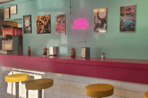 A colorful diner interior with a pink counter, yellow stools, and retro posters on a turquoise wall. Neon sign visible.