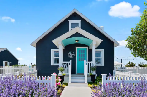 Charming black house with turquoise door, surrounded by colorful flowers and a white picket fence under a blue sky.