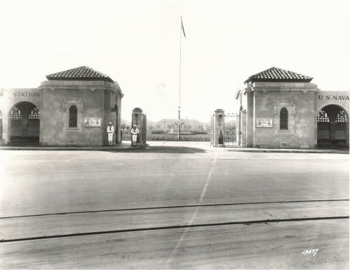 Historic entrance to a naval station, featuring two guards and a flag, with buildings visible in the background.