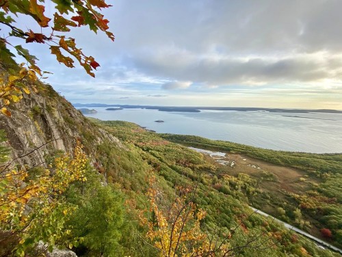 A scenic view of a coastline with autumn foliage, rocky cliffs, and calm waters under a cloudy sky.