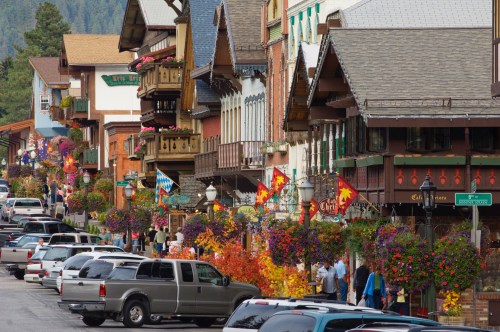 A charming street lined with colorful buildings, flower baskets, and parked cars, set against a backdrop of mountains.