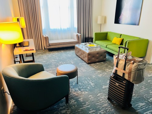 A modern hotel room featuring a green sofa, armchairs, a coffee table, and a suitcase on the floor.