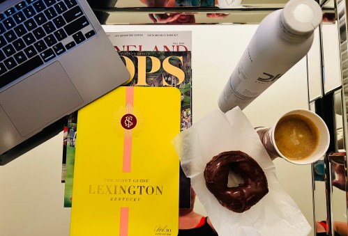 A laptop, a magazine, a yellow guide to Lexington, a coffee cup, a donut, and a water bottle on a reflective surface.