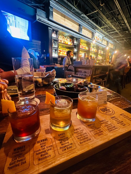 A wooden flight board with three cocktails, garnished with fruit, on a table in a lively bar setting.
