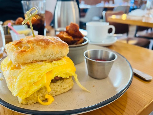 A breakfast plate featuring a biscuit sandwich with egg and cheese, served with a side of potatoes and a small cup of ketchup.