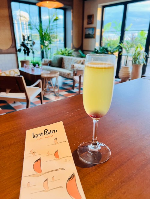 A glass of yellow drink on a wooden table with a menu, surrounded by a cozy, plant-filled café interior.