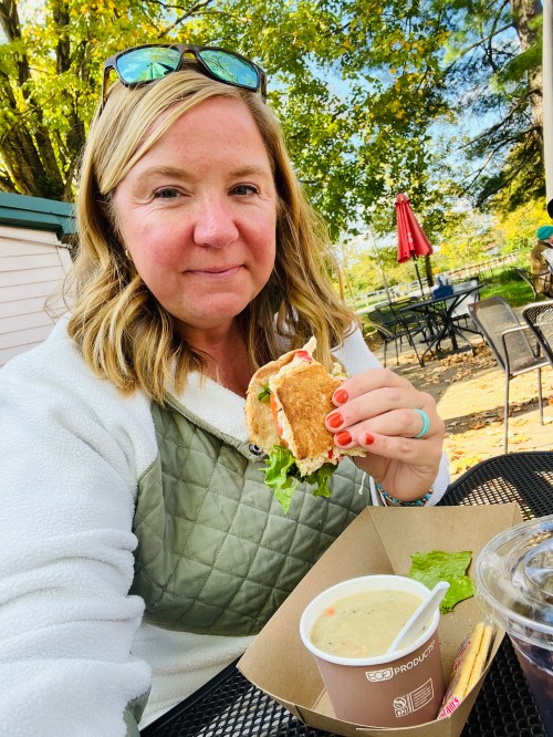 A woman with blonde hair holds a sandwich while sitting outdoors at a table, enjoying a meal on a sunny day.