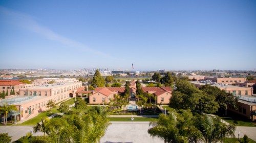 A panoramic view of a campus with buildings, palm trees, and a distant city skyline under a clear blue sky.