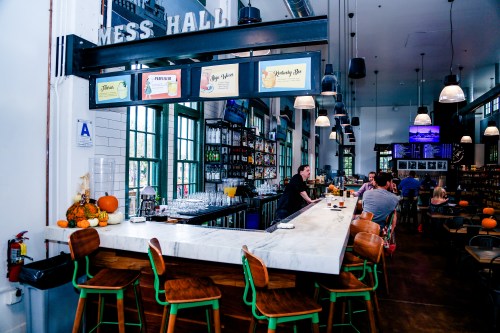A modern bar area in a spacious hall, featuring a marble counter, shelves of drinks, and patrons enjoying the atmosphere.
