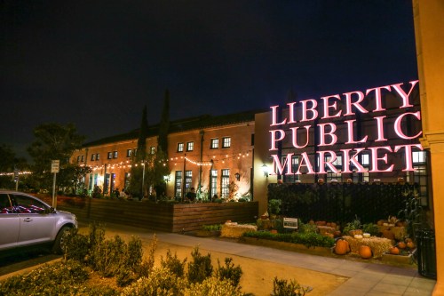 Night view of Liberty Public Market, featuring bright signage and festive lights, surrounded by plants and pumpkins.
