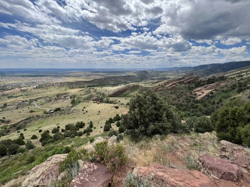 A panoramic view of rolling hills and valleys under a cloudy sky, with rocky formations in the foreground.