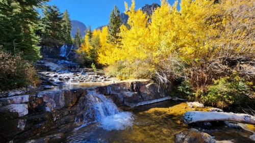 A serene waterfall cascades over rocks, surrounded by vibrant autumn foliage and clear blue skies.
