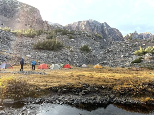 Two hikers stand near colorful tents in a mountainous landscape with rocky terrain and a small pond.