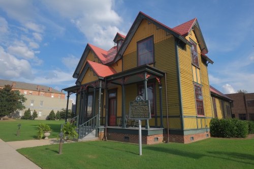 A colorful Victorian-style house with a red roof, surrounded by green grass and a historical marker in front.