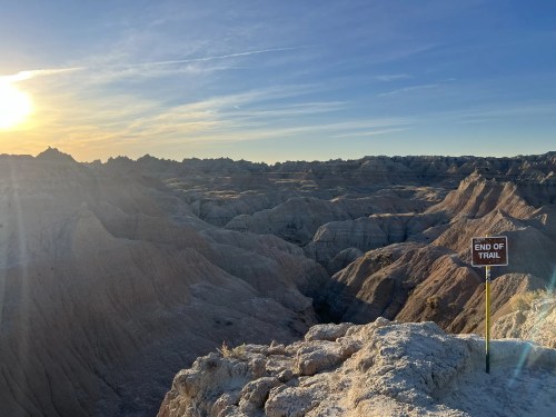 A scenic view of rugged terrain at sunset, featuring a sign that reads "END OF TRAIL" on a rocky ledge.