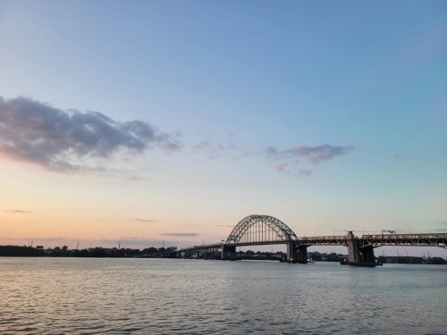 A bridge spans a calm river at sunset, with a clear sky and soft clouds in the background.