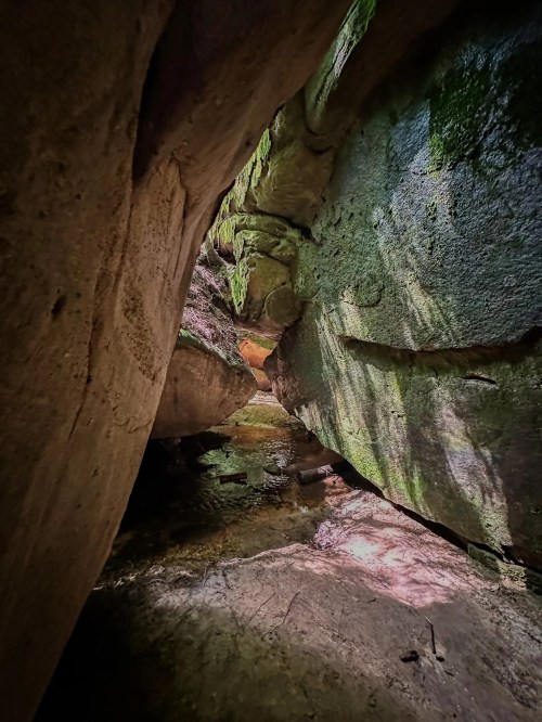 A narrow passageway between moss-covered rocks, leading to a light-filled area with a small stream.