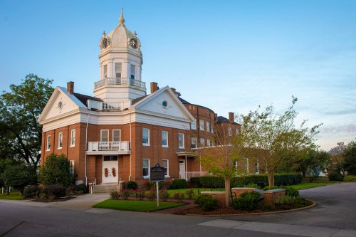 Historic brick building with a clock tower, surrounded by trees and landscaped gardens, under a clear blue sky.