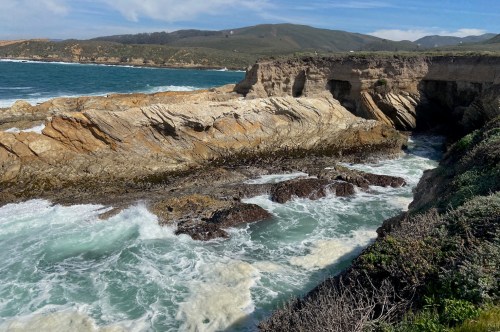 Rocky coastline with crashing waves, lush greenery, and distant hills under a partly cloudy sky.