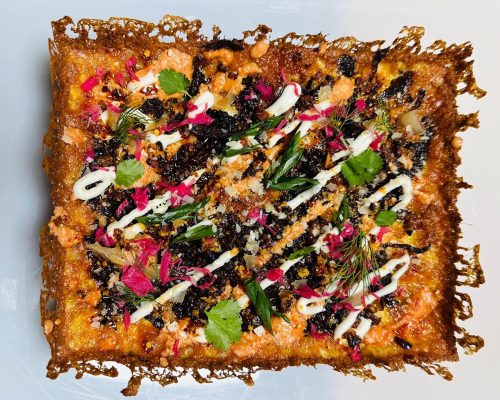 A vibrant, rectangular dish topped with colorful herbs, edible flowers, and a crispy edge, showcasing a mix of textures.