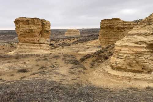 Rock formations rise from a barren landscape under a cloudy sky, with sparse vegetation and dry terrain surrounding them.