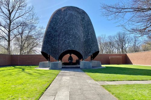 A unique, curved structure with a shingled roof, surrounded by grass and trees, set against a clear blue sky.