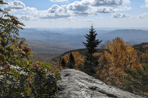 A scenic view of mountains and valleys in autumn, with colorful foliage and a rocky foreground under a cloudy sky.
