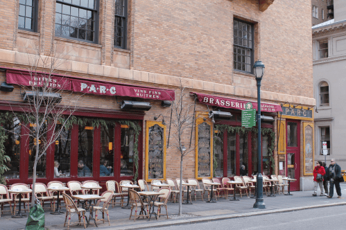 A cozy restaurant exterior with outdoor seating, featuring red awnings and a brick facade. People walk by on the street.