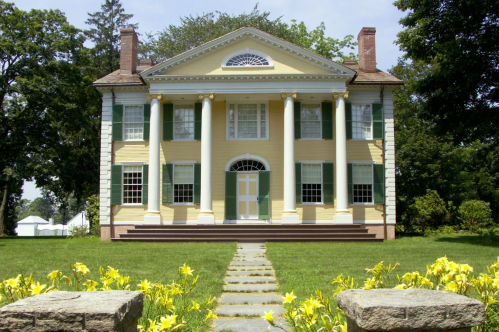 A historic yellow mansion with white columns, green shutters, and a flower-lined pathway leading to the entrance.