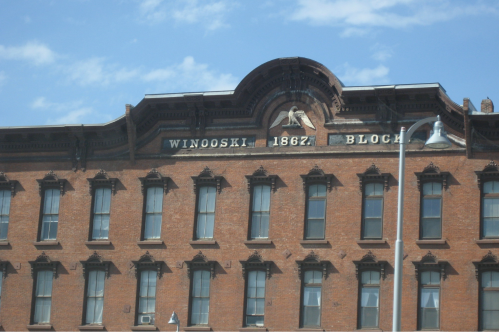 Historic brick building with "Winooski 1867 Block" inscribed, featuring decorative architectural details and a blue sky.