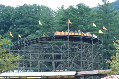 A wooden roller coaster with yellow cars climbs a hill, surrounded by green trees and colorful flags.
