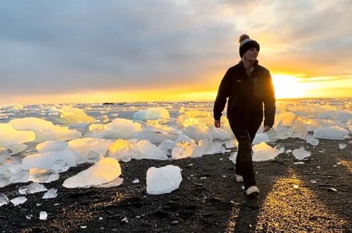 A person walks on a black sand beach surrounded by ice chunks, with a golden sunset in the background.