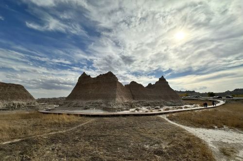 A winding path leads through rugged, layered rock formations under a cloudy sky with the sun peeking through.