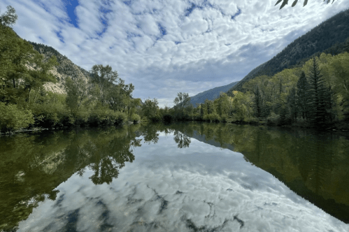 A serene river reflects a cloudy sky and surrounding trees, framed by mountains in the background.