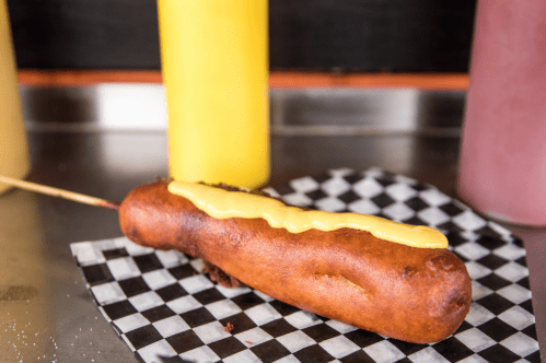 A golden-brown corn dog on a checkered paper, topped with yellow mustard, with condiment bottles in the background.