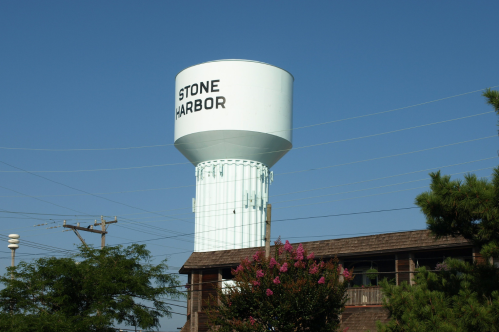 A light blue water tower labeled "STONE HARBOR" stands against a clear blue sky, surrounded by trees and power lines.
