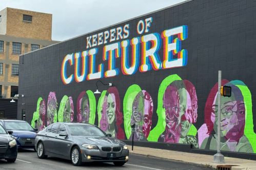 A vibrant mural titled "Keepers of Culture" featuring colorful portraits of diverse individuals against a dark background.
