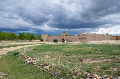 A historic adobe building under a cloudy sky, surrounded by grassy fields and a winding path.